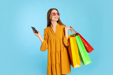 Fototapeta na wymiar Happy young woman with shopping bags and smartphone on light blue background. Big sale