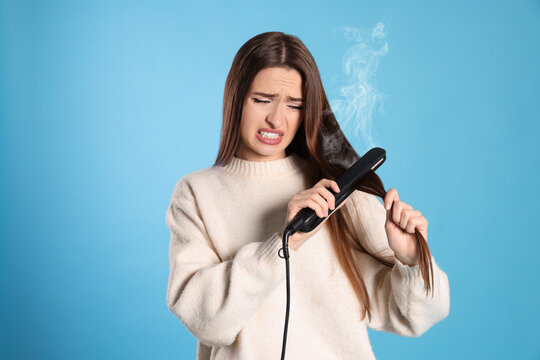 Stressed young woman with flattening iron on light blue background. Hair damage