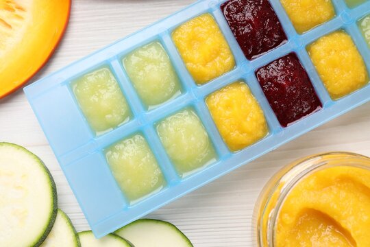 Different purees in ice cube tray and ingredients on white table, flat lay. Ready for freezing
