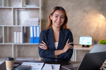 Beautiful Asian businesswoman sitting smiling, cross arms and opened her laptop and happily looking at the camera in the office.