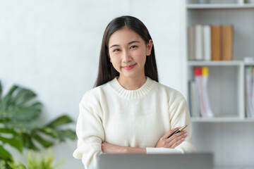 Beautiful Asian businesswoman sitting smiling, cross arms and opened her laptop and happily looking at the camera in the office.