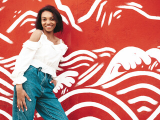 Portrait of young beautiful black woman. Smiling hipster model in white clothes. Sexy carefree female posing on the street background near wall. Cheerful and happy outdoors. At sunset. In sunglasses
