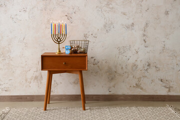 Basket with bells, dreidels and menorah for Hanukkah on table near light wall