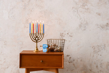Basket with bells, dreidels and menorah for Hanukkah on table near light wall