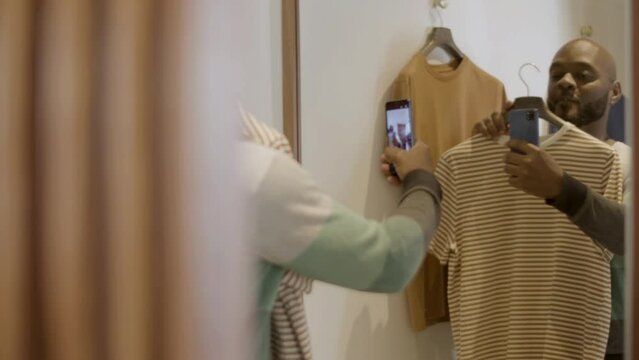 Happy black guy taking selfie in dressing room holding hanger while trying on T-shirt. Handsome African American man taking picture with phone in front of mirror. Modern technology, shopping concept.