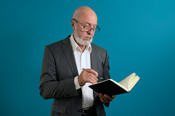 An elderly man in a suit with a notebook and a pen is thinking about a task. on a blue background