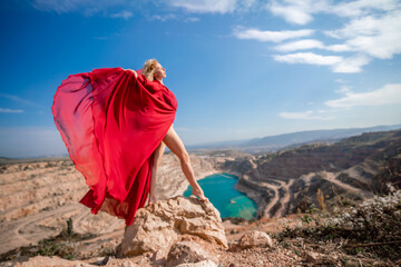 Side view of a beautiful sensual woman in a red long dress posing on a rock high above the lake in the afternoon. Against the background of the blue sky and the lake in the form of a heart