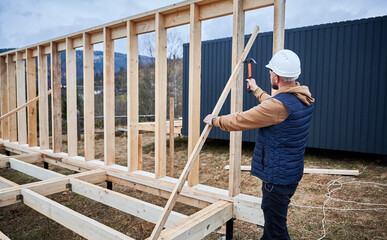 Fototapeta premium Man worker building wooden frame house on pile foundation. Carpenter hammering nail into wooden truss, using hammer. Carpentry concept.