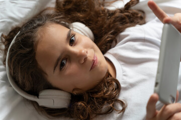 beautiful teenage girl looks at smartphone screen with headphones in bed in the morning. Happy adult child wakes up and listens to music, plays games, studies with lessons on the phone