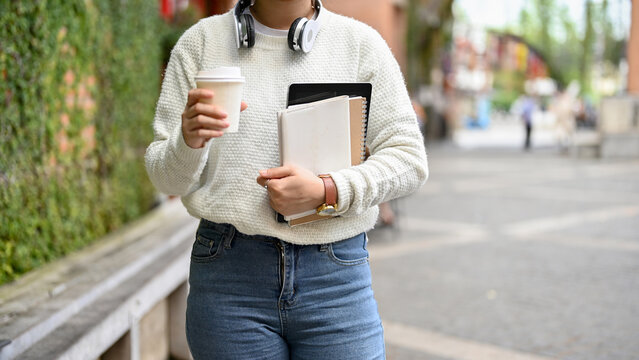 Female college student walking in a university street while holding textbooks and coffee cup