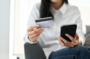 A young Asian woman holding a credit card and her smartphone while relaxes sitting on sofa.