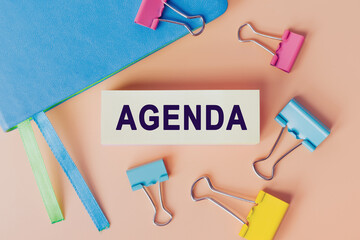 Text AGENDA written on white paper. Note near notebook and many colourful paper clips, top view. Business concept. Home Office