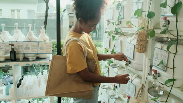 Black female customer fills liquid soap in recycle bottle from reusable containers in zero-waste and refill store, environment-friendly shop. Thai text labels mean shampoo, soap, and home appliances.