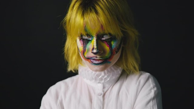 Young woman with colorful face art posing ob black background, clown makeup
