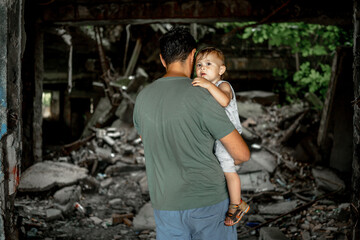 The father holds the child in his arms. In front of them is a city destroyed after the war.