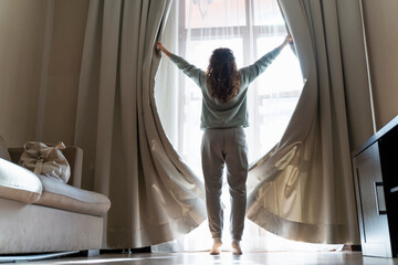Happy young woman opens the curtains and looks out the window. Beautiful brunette opens the curtains to let the morning sunlight