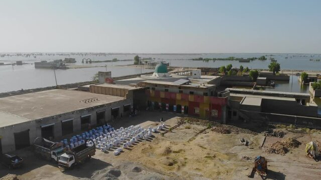 Aerial Dolly Back Shot Of Row Of White Food Sacks In Courtyard Beside Flooded Landscape In Jacobabad. Pedestal Up
