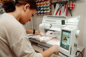 A woman tailor makes embroidery on an embroidery machine in a home workshop.Mending clothes concept,small business.Selective focus,close-up. - obrazy, fototapety, plakaty