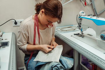A woman mends jeans, sews a patch on a hole in the home studio.Mending clothes concept,reusing old jeans.Small business. - obrazy, fototapety, plakaty