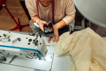 Caucasian woman sews on a sewing machine in a home workshop and records the broadcast on a smartphone,hands close-up.Small business.Blogging concept.Selective focus.