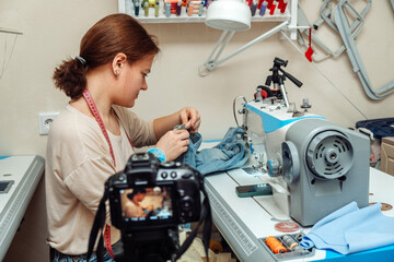 A woman mends jeans, sews a patch on a hole in the home studio and records the broadcast on a camera.Mending clothes concept,reusing old jeans.Small business.Blogging concept. - obrazy, fototapety, plakaty