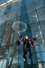 Cheerful happy young girl jumps with transparent umbrella against glass facade of building. Bottom view. Vertical frame.
