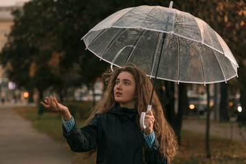 Girl with brown hair walks in fall park. Young woman standing under transparent umbrella to see if it is raining