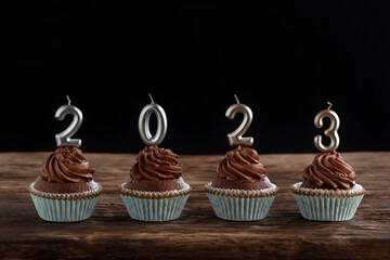 Chocolate cupcakes with buttercream frosting with silver 2023 candles on wooden table on black background.