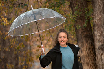 Student in black coat and with transparent umbrella poses for photo. Girl with dark hair walks in autumn park