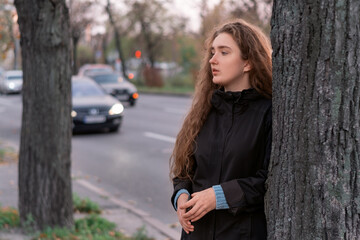 Young brooding woman stands by tree near highway on cloudy autumn day. Teen girl in coat on road...