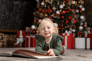 Portrait of cute little blonde girl lying on floor with large book under Christmas tree . Children and New Year mood