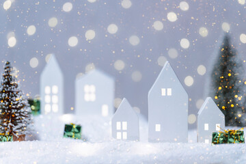 Christmas cute little houses with tree in snow. Glowing festive garland on light background. Cozy fairytale atmosphere with home decor with selective focus in the foreground New Year card.