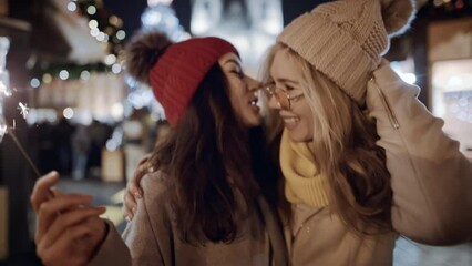 Two female friends meet at xmas party outdoors at Old decorated town square. Happy time together - Asian girl making surprise for her mate. Celebrating New Year holidays vacation. Christmas joy
