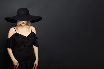 Fototapeta na wymiar Stylish woman wearing black dress and black broad brim wide hat on black background