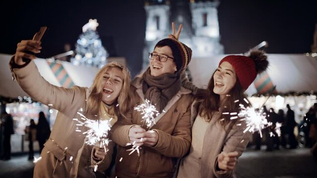 Celebrating New Year holidays vacation. Happy dancing three friends with sparklers smile at decorated town square. Xmas Christmas winter night atmosphere. Excited people take selfie on smartphone
