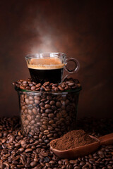 a glass jar filled with roasted coffee beans, on top of a steaming cup of espresso.