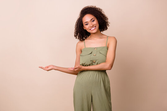 Photo of gorgeous pretty lovely woman with curly hairdo wear khaki overall demonstrating empty space isolated on beige color background