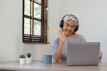 Smiling senior asian woman with headphones on her head sitting at a table in front of a laptop and...
