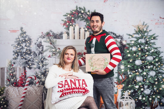 Couple in Christmas outfits jumper dress festive standing sitting posing with cushion and gift box Christmas decoration background white dress bright studio picture with glitter light leak