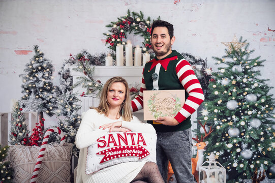 Couple in Christmas outfits jumper dress festive standing sitting posing with cushion and gift box Christmas decoration background white dress bright studio picture 