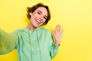 Photo portrait of cute young woman video call streaming waving hand say hello wear trendy green look isolated on yellow color background