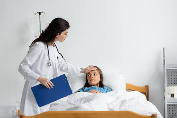 Fototapeta premium Doctor with clipboard examining forehead of child in patient gown on hospital bed.