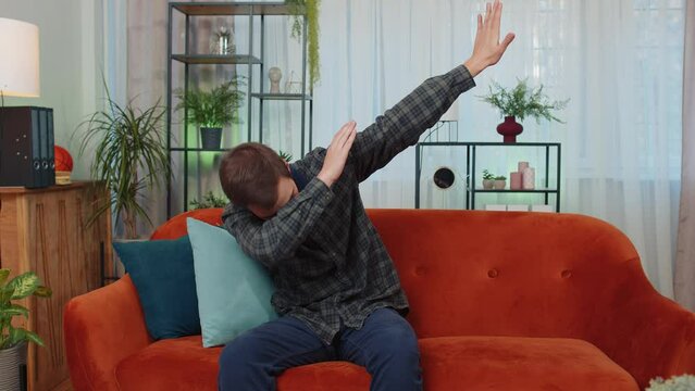 Trendy cheerful positive caucasian man having fun dancing and moving to rhythm, dabbing raising hands, making dub dance gesture at modern home apartment. Young guy in living room on couch. Lifestyle