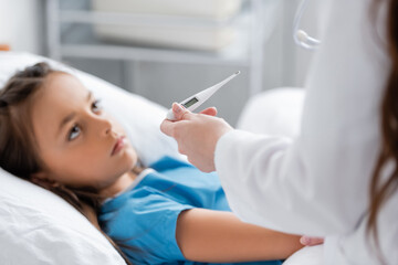 Naklejka premium Doctor in white coat holding electronic thermometer near blurred child in hospital ward.