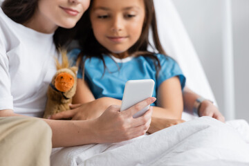 Woman using smartphone near daughter with soft toy on bed in hospital.