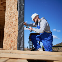 Male builder installing black corrugated iron sheet used as facade of future cottage, blue sky on...