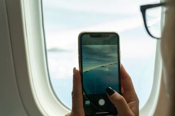 Hands of unrecognizable woman with glasses taking a picture of the clouds and the wing of the plane through the round window.