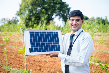 Confident banking officer showing solar panel by looking at camera at agricultural land - concept...