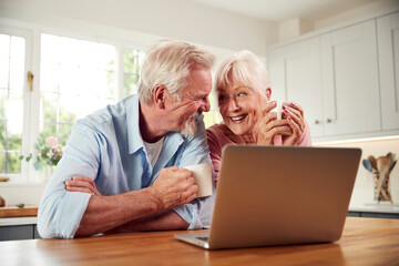 Retired Senior Couple Sitting In Kitchen At Home Drinking Coffee And Using Laptop