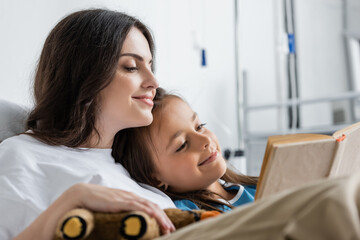 Positive woman holding soft toy while reading book with mother in hospital ward.